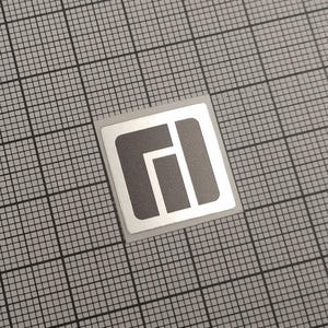 May include: A square sticker with a gray geometric design on a white background. The sticker is centered on a gray grid background. The geometric design consists of a stylized letter "I" and a stylized letter "L".