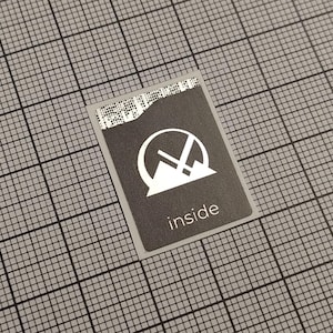 Pode incluir: Um pequeno autocolante retangular cinzento escuro com um gráfico branco e a palavra "inside". O gráfico é um design estilizado de montanha e seta dentro de um círculo. O autocolante está sobre um fundo de grelha cinzenta.