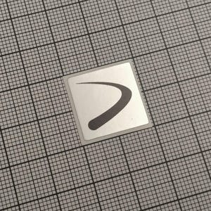 May include: A small, square sticker with a silver border. The sticker features a dark gray, curved design resembling a checkmark or a stylized swoosh, set against a white background. The sticker is placed on a gray grid.