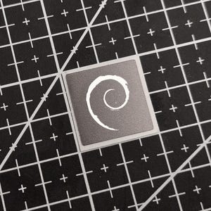 Puede incluir: Una pegatina cuadrada con fondo gris y diseño de espiral blanco. La pegatina está sobre una esterilla de corte negra con líneas de cuadrícula blancas.