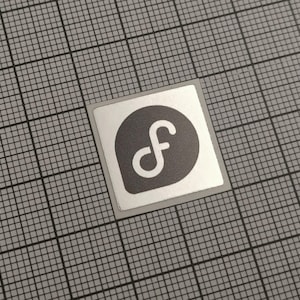 Op de afbeelding: Een vierkante sticker met een zilveren rand. De sticker heeft een donkergrijze cirkel met een witte gestileerde letter "f" erin. De sticker is geplaatst op een grijze rasterachtergrond.