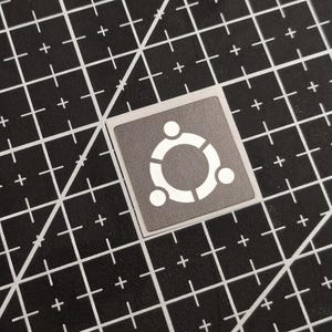 Op de afbeelding: Een vierkante sticker met een grijze achtergrond en een witte afbeelding van een cirkel met drie verbonden segmenten en drie stippen. De sticker ligt op een zwarte snijmat met witte rasterlijnen.