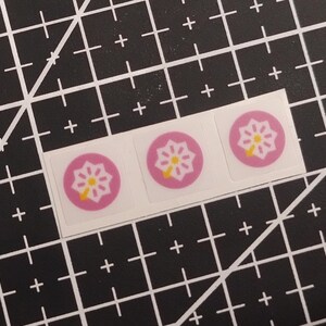 May include: Three square stickers arranged in a horizontal row, each featuring a circular pink background with a white eight-pointed star design and a small yellow arrow pointing toward the bottom left corner of the circle.