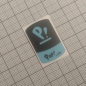 May include: A rectangular sticker with a black top and a light blue bottom. The sticker features the word "Pop!_os" in light blue, along with a stylized "P!" symbol in the same color. The sticker is on a grid background.
