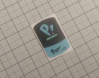 Puede incluir: Una pegatina rectangular con la parte superior negra y la inferior azul claro. La pegatina presenta la palabra "Pop!_os" en azul claro, junto con un símbolo "P!" estilizado del mismo color. La pegatina está sobre un fondo de cuadrícula.