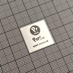 Puede incluir: Una pegatina cuadrada con el logotipo de Pop!_os, una 'P' estilizada dentro de un círculo con un signo de exclamación. El texto 'Pop!_os' está debajo, con 'open source' debajo. La pegatina está sobre un fondo de cuadrícula gris.