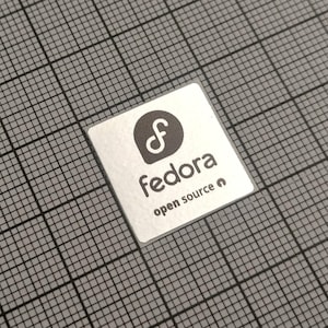 May include: A square, silver sticker with the word "fedora" in a sans-serif font, and the words "open source" below it. The sticker has a stylized "f" logo. The sticker is on a gray grid background.