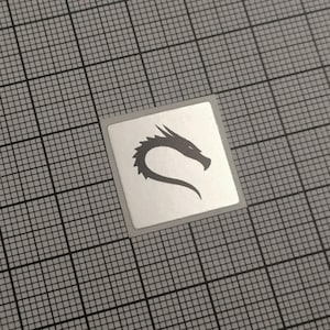 Op de afbeelding: Een vierkante sticker met een zilveren achtergrond en een donkergrijze draak silhouet. De kop van de draak is naar rechts gericht, met een gebogen lichaam. De sticker ligt op een grijze rasterachtergrond.