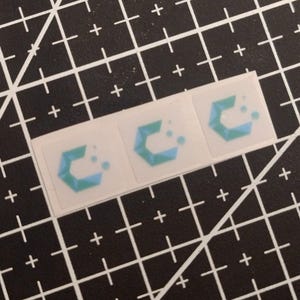 May include: Three square stickers arranged in a horizontal row, each featuring a geometric logo design in shades of blue and teal. The logo consists of a stylised letter C composed of triangular segments with two small circles to the right.