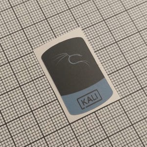 May include: A sticker featuring a black rectangle with a white stylized scorpion design and a light blue bottom section. The word "KALI" is printed in a white box on the blue section. The sticker is on a grid background.