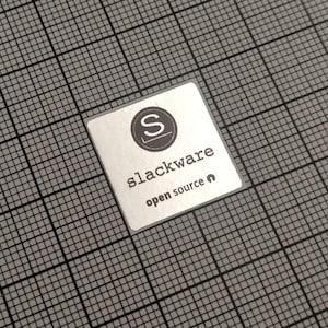 Peut inclure: Un autocollant carré avec le texte "slackware open source" en police sans-serif. L'autocollant a un fond argenté et un logo noir et blanc avec la lettre "S" dans un cercle. L'autocollant est sur une grille grise.