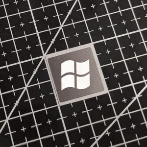 May include: A square sticker with the Windows logo, a white design of four curved panes within a dark grey square. The sticker is on a black cutting mat with a white grid pattern.