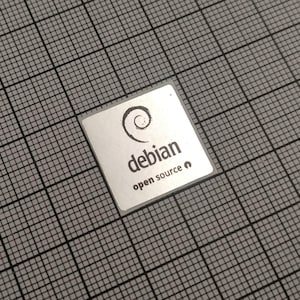 Könnte beinhalten: Ein kleines, quadratisches, silberfarbenes Objekt mit dem Wort "debian" und einem Spiral-Logo in Schwarz aufgedruckt. Unter dem Wort "debian" steht der Text "open source". Das Objekt befindet sich vor einem grauen Gitterhintergrund.