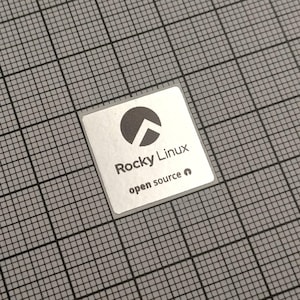 May include: A square, silver sticker with the words "Rocky Linux" and "open source" printed in black. The sticker features a black graphic of a circle with a white arrow. The sticker is on a gray grid.