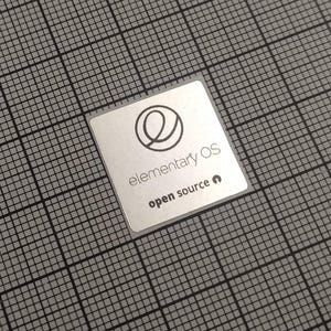 Może przedstawiać: Kwadratowa naklejka z zaokrąglonymi rogami, z logo i tekstem "elementary OS". Logo to stylizowane "e" w okręgu. Tekst "open source" znajduje się poniżej. Naklejka znajduje się na szarym tle w kratkę.