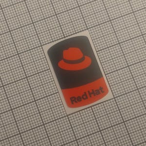 Può includere: Un adesivo rettangolare con un logo a cappello rosso su sfondo nero. La sezione inferiore è rossa con la scritta bianca "Red Hat". L'adesivo è su uno sfondo a griglia.