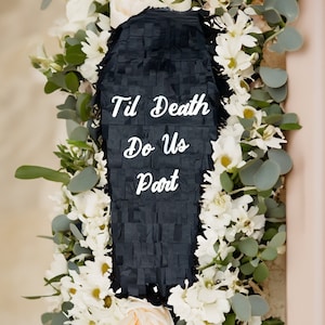 May include: A black coffin-shaped piñata with the text "Til Death Do Us Part" in white script, surrounded by a wreath of white flowers and green foliage. The floral arrangement includes roses and eucalyptus.