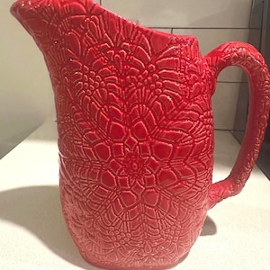 Pode incluir: Uma jarra de cerâmica vermelha vibrante com um padrão floral texturizado. A jarra tem um bico curvo e uma alça grande e arqueada. O design é em relevo, criando uma superfície tátil. Esta jarra decorativa é adequada para servir bebidas.