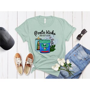 May include: A light green t-shirt with the text "Ponte Kicks and Keep Going" and a cartoon graphic of a candle and a jar with a face. The shirt is styled with jeans, sunglasses, and sneakers.