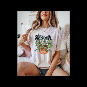 Peut inclure: T-shirt beige avec l'inscription "La Señora De las Plantas" et un motif de plante en pot avec des feuilles vertes. Le design est en noir, vert et orange. Le t-shirt est porté.
