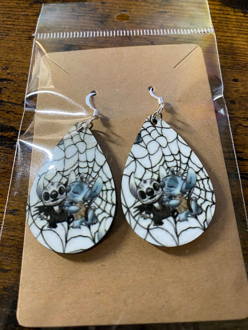 K&ouml;nnte beinhalten: Tropfenf&ouml;rmige Ohrringe mit einem Cartoon-Charakter-Design. Die Ohrringe haben einen wei&szlig;en Hintergrund mit einem schwarzen Spinnennetz-Muster. Der Cartoon-Charakter befindet sich in der Mitte der Ohrringe. Die Ohrringe befinden sich in einer durchsichtigen Plastikt&uuml;te.