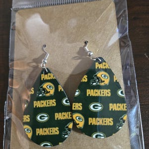 May include: Teardrop-shaped earrings featuring a green and gold pattern with the words "PACKERS" and a football helmet logo. The earrings are suspended from silver-colored fish hook ear wires. The earrings are displayed on a brown card inside a clear plastic bag.