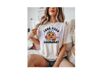 Puede incluir: Camiseta beige claro con un diseño gráfico colorido de una calabaza sonriente sosteniendo una taza. El texto "¿Qué pasa calabaza?" está encima y debajo del diseño de la calabaza. La camiseta la lleva una persona.
