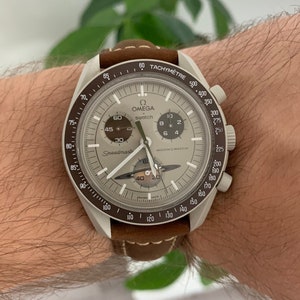 Può includere: Un orologio da polso cronografo argento e marrone con cinturino in pelle marrone. Il quadrante dell'orologio presenta una scala tachimetrica, un indicatore di fase lunare e il testo "Speedmaster" e "Moon's Watch".