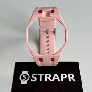 Puede incluir: Una caja y correa de reloj de silicona rosa claro con un marco octogonal geométrico y pequeños detalles rojos en los puntos de unión. Debajo hay una tarjeta rectangular negra con el texto blanco STRAPR y la dirección web straprwatch.com.