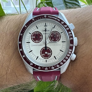 May include: A white and red Omega Speedmaster MoonSwatch watch with a burgundy strap. The watch face features a tachymeter scale, chronograph subdials, and the words "Speedmaster" and "MoonSwatch".