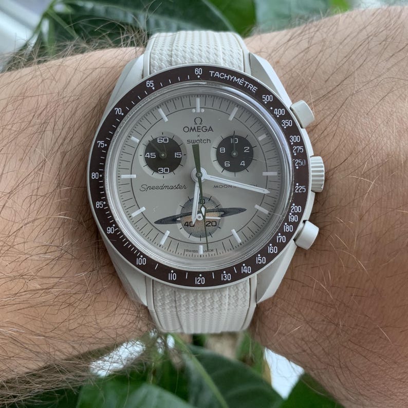 May include: A white Omega Swatch Speedmaster watch with a light gray face and a white strap. The watch features a tachymeter bezel and multiple sub-dials. The watch face displays the text "OMEGA swatch Speedmaster".