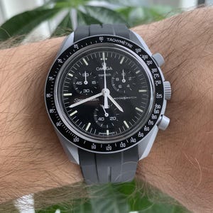 May include: A black chronograph wristwatch with a silver band and a black rubber strap. The watch face has white markings and the words "Omega swatch" and "Moonswatch" printed on it.