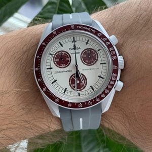 May include: A white and red Omega Speedmaster MoonSwatch watch with a grey silicone strap. The watch face features a chronograph with three subdials and the text "Speedmaster" and "MoonSwatch".