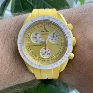 May include: A yellow Omega Speedmaster MoonSwatch watch with a white face and red accents. The watch has a tachymeter scale around the edge of the face and a chronograph function.