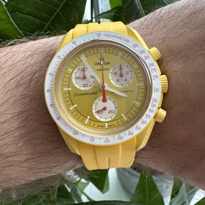 May include: A yellow Omega Speedmaster chronograph wristwatch with a white tachymeter bezel and red markings. The watch face is yellow with black and white markings and the words "Omega swatch Speedmaster" printed on it.