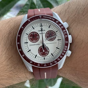 May include: A white and silver chronograph wristwatch with a red and white silicone strap. The watch face has a white background with black markings and the words "Speedmaster" and "Moonswatch" printed on it. The watch also has a tachymeter scale around the outer edge of the face.