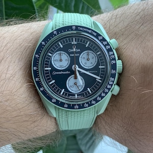 May include: A light green Omega Speedmaster watch with a black dial and white hands. The watch features a tachymeter bezel and three sub-dials. The watch is on a light green strap.