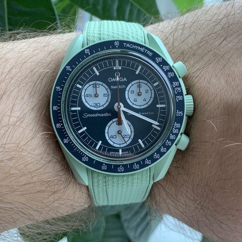 May include: A light green Omega Speedmaster watch with a black dial and white hands. The watch features a tachymeter bezel and three sub-dials. The watch is on a light green strap.