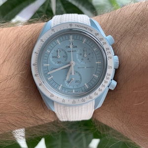 May include: A light blue and white wristwatch with the text "OMEGA" and "Moon Watch" on the face. The watch has a white strap and a light blue bezel. The watch also has a tachymeter.