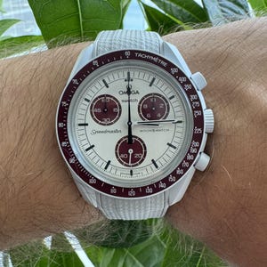 May include: A white Omega Speedmaster MoonSwatch watch with a white strap. The watch face is white with burgundy accents and sub-dials. The watch has a tachymeter bezel and the text "Speedmaster" is visible.