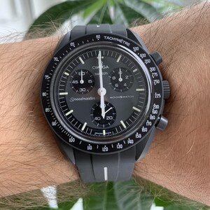 May include: A black Omega Speedmaster MoonSwatch watch with a black face and white markings. The watch has a tachymeter scale on the bezel and a date window at the 3 o'clock position.