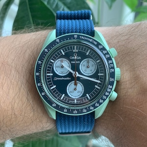 Op de afbeelding: Een mintgroen Omega Speedmaster chronograafhorloge met een zwarte wijzerplaat en een blauwe nylon band. Het horloge heeft een tachymeterschaal rond de bezel en een datumvenster op 3 uur.