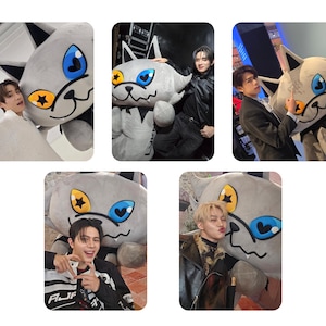May include: Multiple images of a large, gray cat plushie with blue and yellow accents. The plushie has a cartoonish design and is being held by different people in various poses. The cat's eyes are blue and the star-shaped accents are yellow.