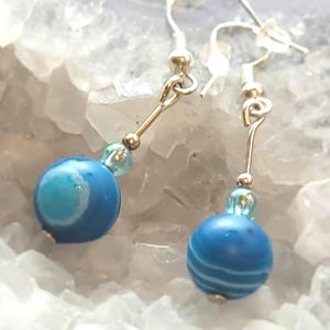 Op de afbeelding: Een paar zilveren oorhangers met blauwe agaat kralen. De kralen hebben een wit swirl-patroon.