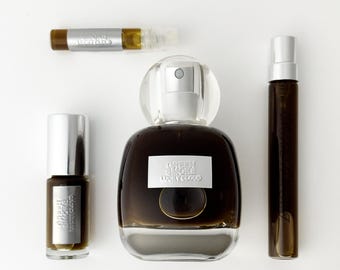 Può includere: Una collezione di flaconi di profumo che include una grande bottiglia di vetro arrotondata e tre fiale cilindriche più piccole. Ogni contenitore è riempito con un liquido marrone scuro e presenta un'etichetta argentata con il testo Green Smoke Lucky Cloud.