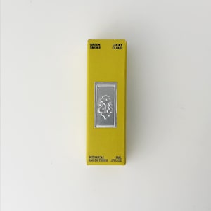 K&ouml;nnte beinhalten: Eine rechteckige gelbe Schachtel mit schwarzem Text, der Green Smoke Lucky Cloud und Botanical Eau de Terre 5ml lautet. Ein mittiges silbernes rechteckiges Emblem zeigt ein gepr&auml;gtes Blumenmuster auf der Vorderseite.
