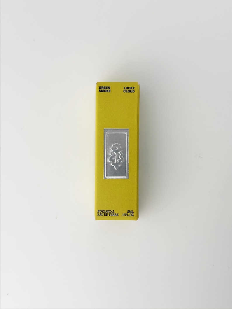 K&ouml;nnte beinhalten: Eine rechteckige gelbe Schachtel mit schwarzem Text, der Green Smoke Lucky Cloud und Botanical Eau de Terre 5ml lautet. Ein mittiges silbernes rechteckiges Emblem zeigt ein gepr&auml;gtes Blumenmuster auf der Vorderseite.