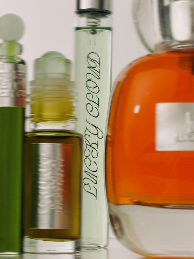 K&ouml;nnte beinhalten: Eine Sammlung von Glasparf&uuml;mflaschen, darunter eine hohe, d&uuml;nne Flasche mit dem in Gr&uuml;n gedruckten Text Lucky Cloud, eine kleine Roll-on-Flasche mit dunkel bernsteinfarbener Fl&uuml;ssigkeit und eine gro&szlig;e, abgerundete Flasche mit leuchtend orangefarbener Fl&uuml;ssigkeit.