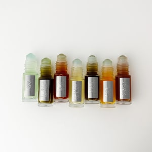 K&ouml;nnte beinhalten: Sieben kleine Glasfl&auml;schchen mit Roll-on-Aufsatz in einer Reihe, die jeweils eine unterschiedlich gef&auml;rbte Fl&uuml;ssigkeit von hellgr&uuml;n bis dunkel bernsteinfarben enthalten. Jede Flasche hat ein silbernes rechteckiges Etikett mit Text.