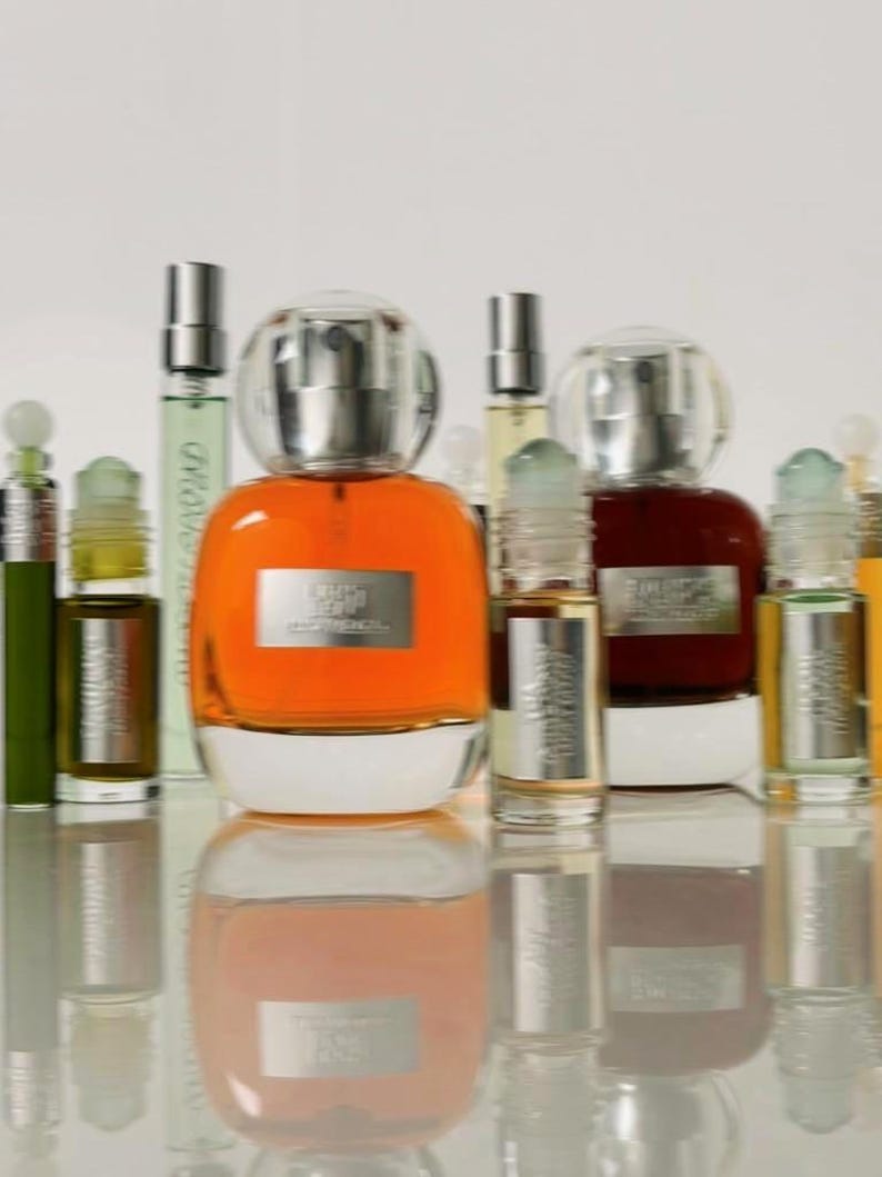 K&ouml;nnte beinhalten: Eine Sammlung von Glasparf&uuml;mflaschen mit silbernen Etiketten und durchsichtigen Verschl&uuml;ssen. Die mittlere Flasche enth&auml;lt eine leuchtend orangefarbene Fl&uuml;ssigkeit, w&auml;hrend andere bernsteinfarbene, gr&uuml;ne und dunkelbraune Fl&uuml;ssigkeiten enthalten.